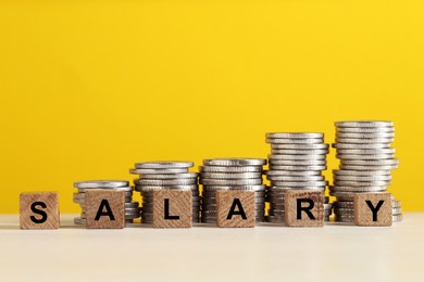 Photo of Word Salary made of cubes and stacked coins on white table against yellow background, closeup