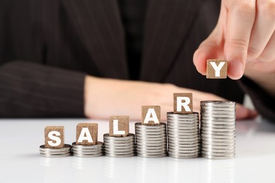 Photo of Woman putting wooden cubes with word Salary and stacks of coins on white table, closeup