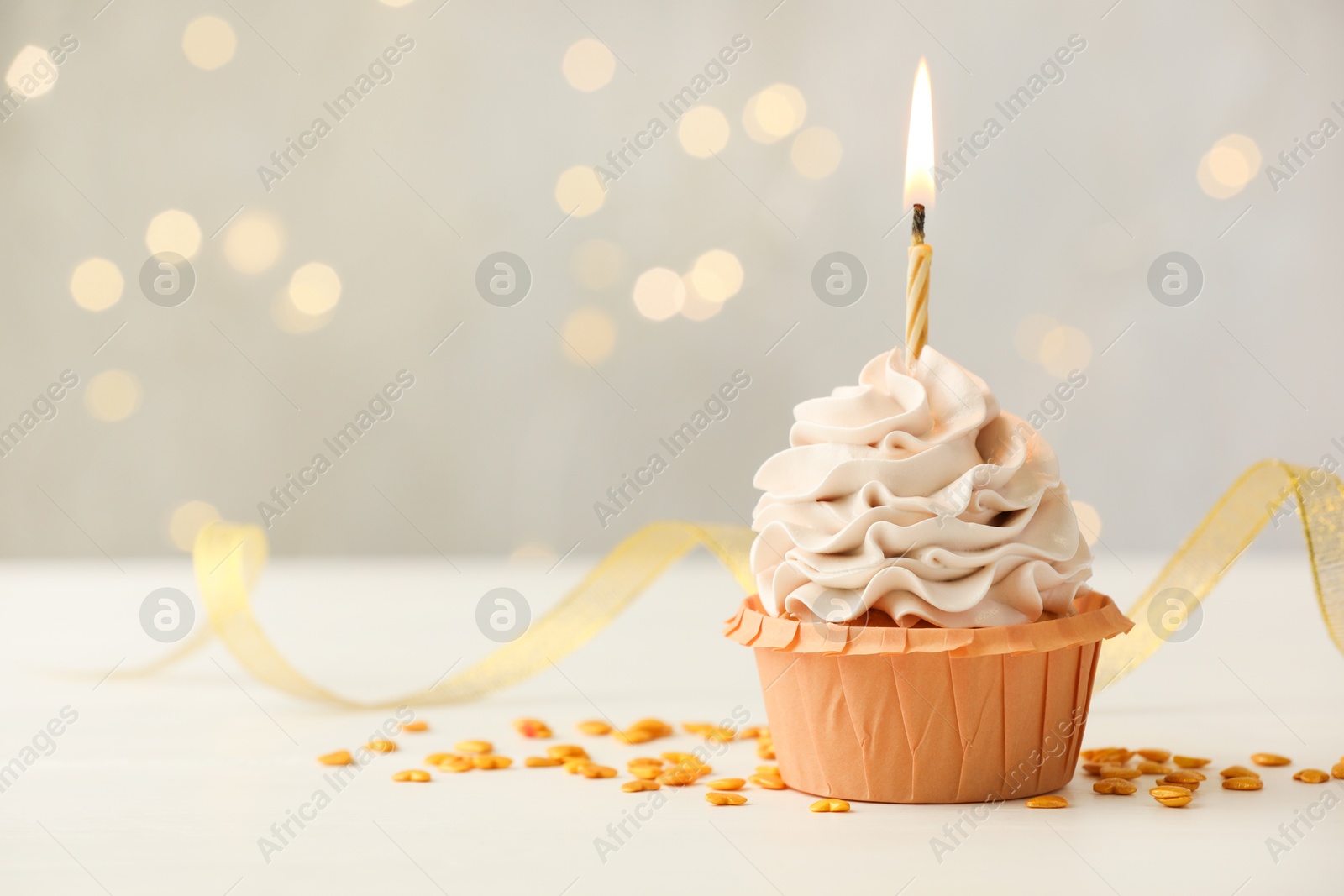 Photo of Birthday cupcake with burning candle on light table against blurred lights. Space for text