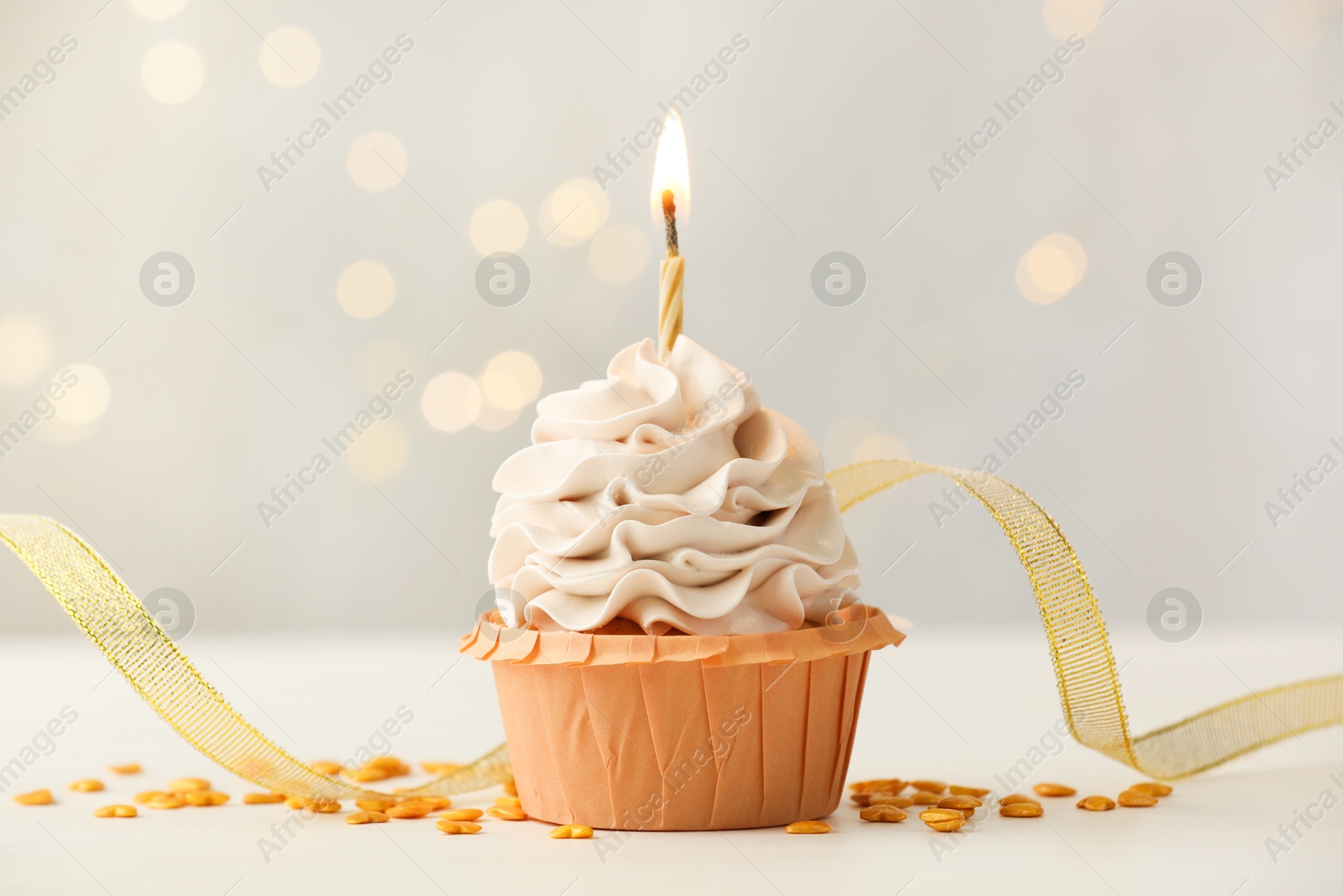 Photo of Birthday cupcake with burning candle on light table against blurred lights