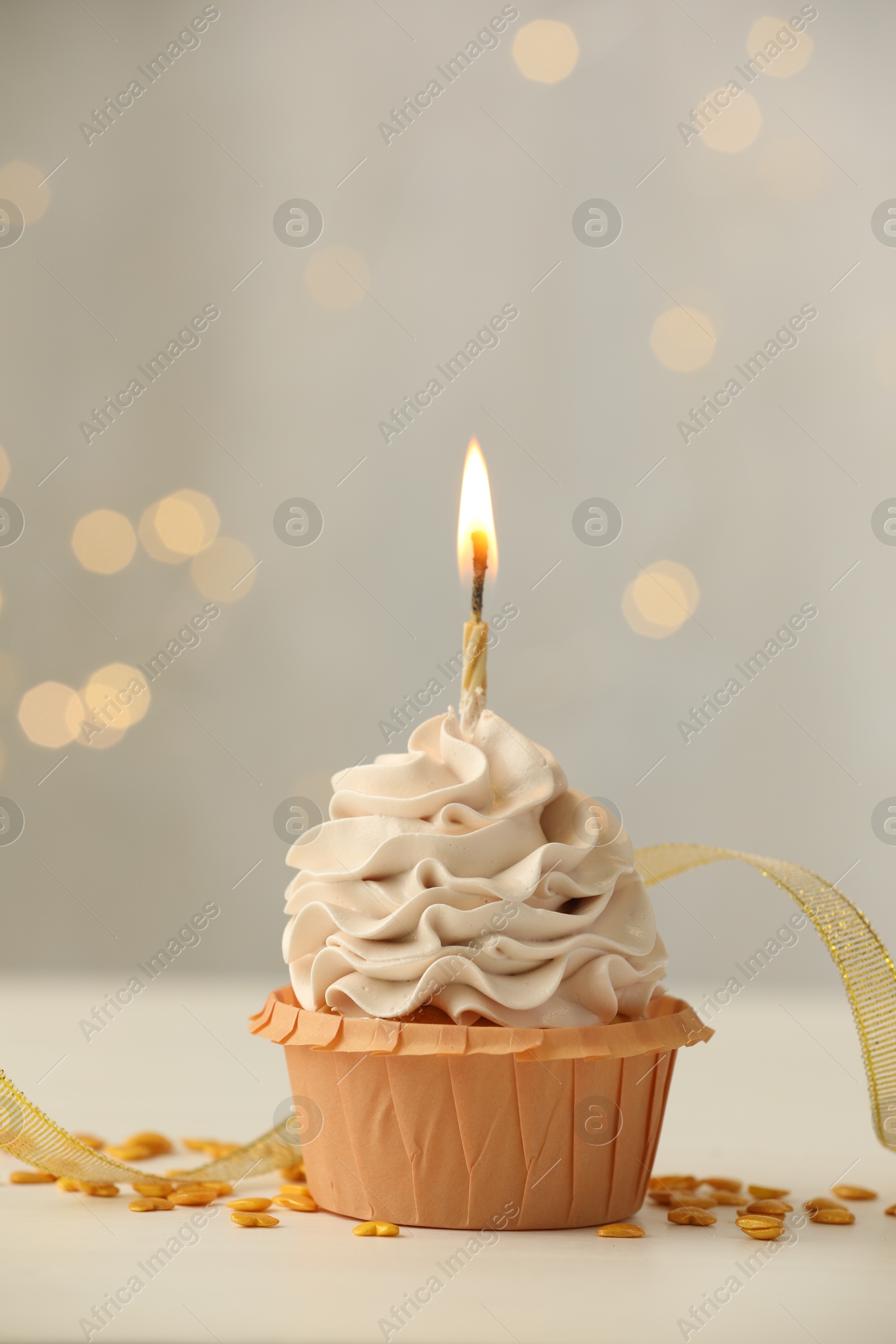 Photo of Birthday cupcake with burning candle on light table against blurred lights