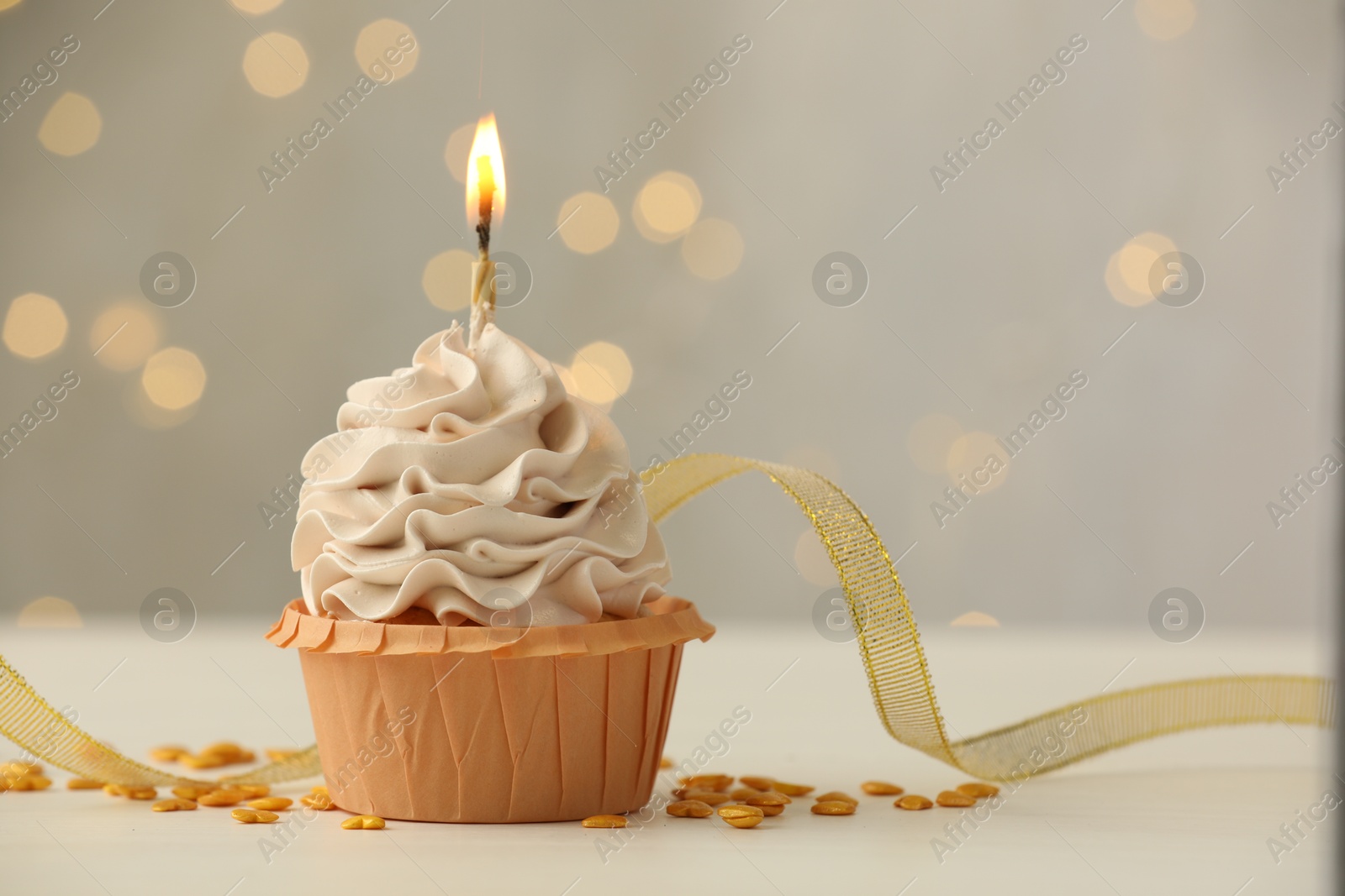 Photo of Birthday cupcake with burning candle on light table against blurred lights. Space for text