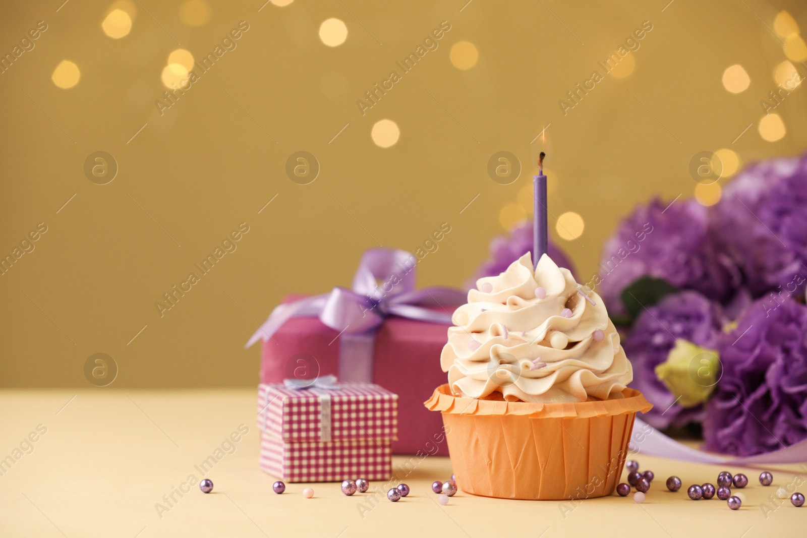 Photo of Birthday cupcake with candle, gifts and flowers on beige table against blurred lights, selective focus. Space for text