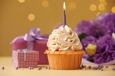 Photo of Birthday cupcake with burning candle, gifts and flowers on beige table against blurred lights, selective focus