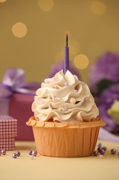 Photo of Birthday cupcake with candle, gifts and flowers on beige table against blurred lights, selective focus