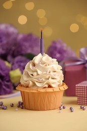 Photo of Birthday cupcake with candle, gifts and flowers on beige table against blurred lights, selective focus