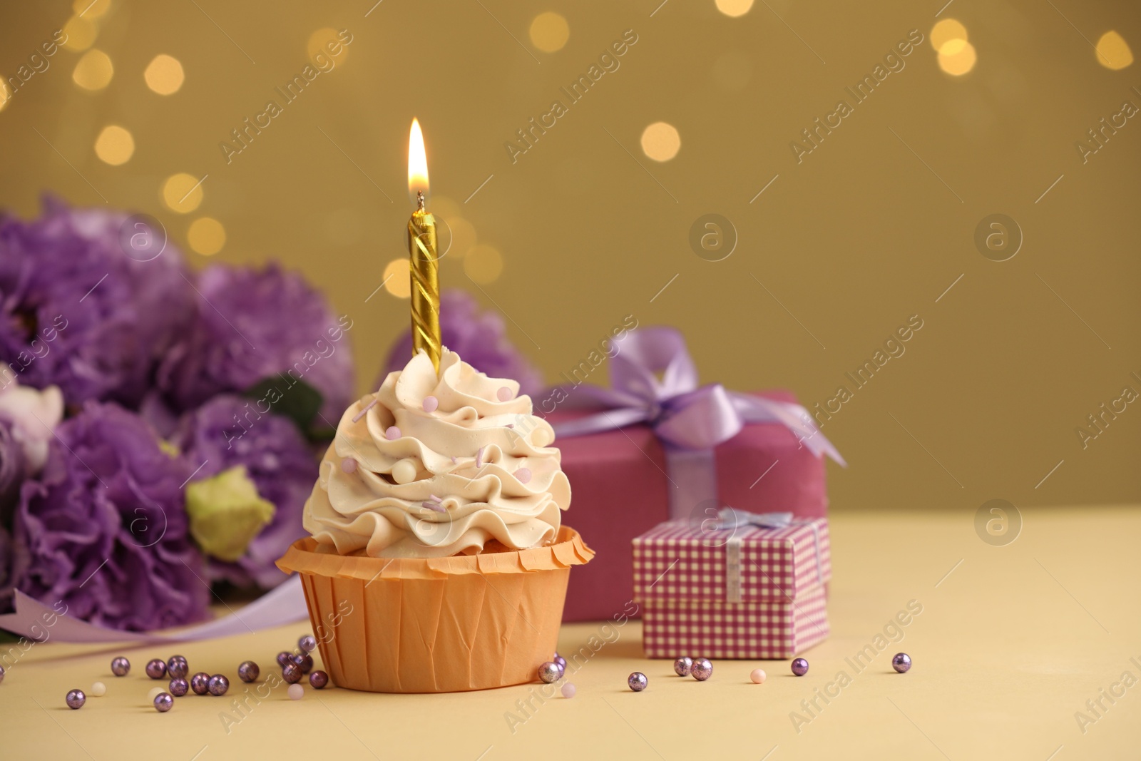 Photo of Birthday cupcake with burning candle, gifts and flowers on beige table against blurred lights, selective focus. Space for text