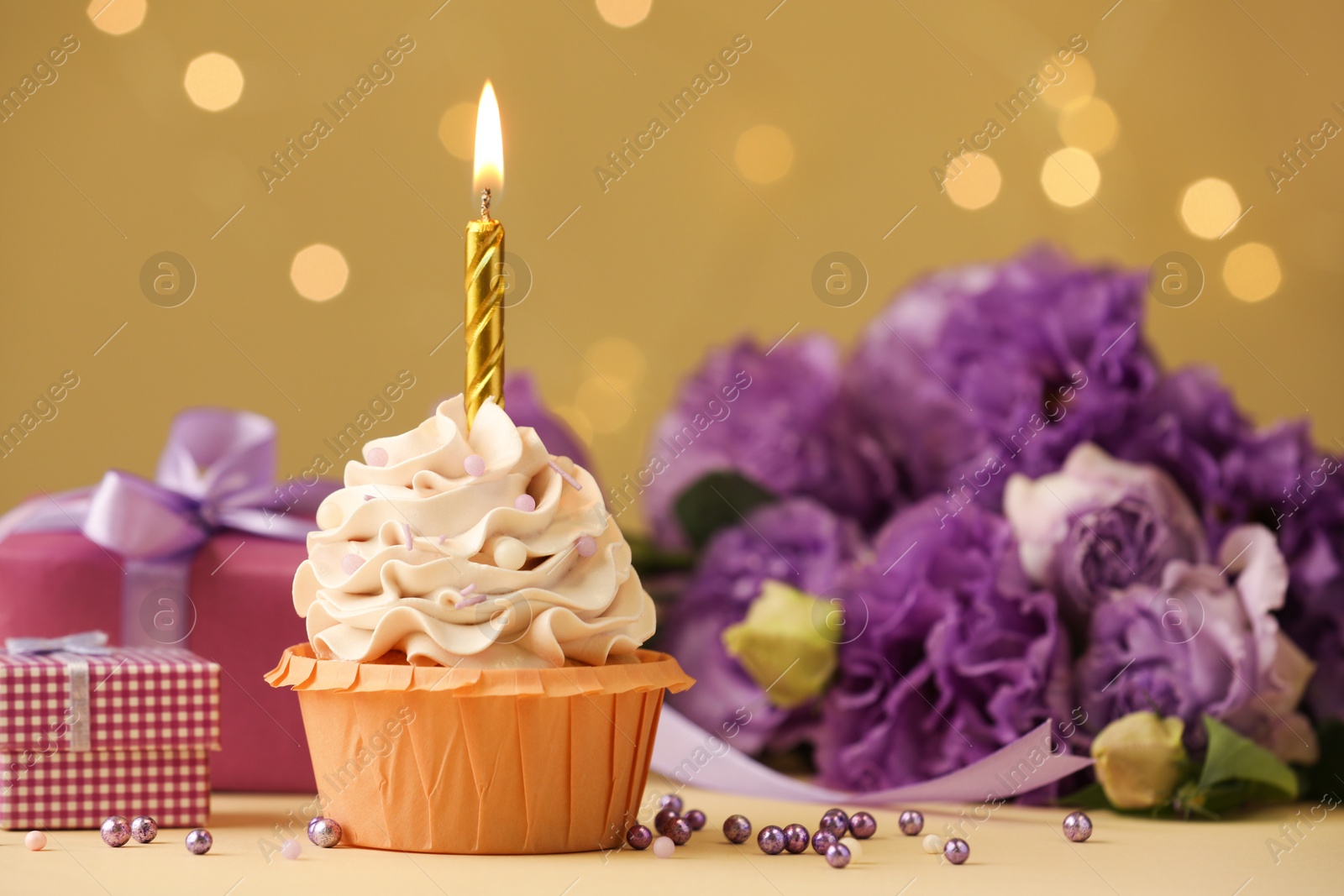 Photo of Birthday cupcake with burning candle, gifts and flowers on beige table against blurred lights, selective focus