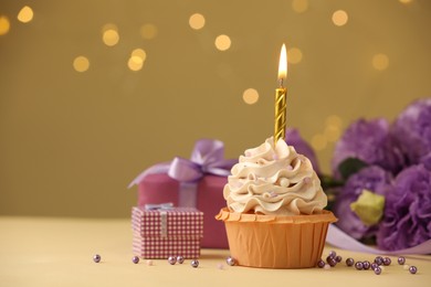 Photo of Birthday cupcake with burning candle, gifts and flowers on beige table against blurred lights, selective focus. Space for text