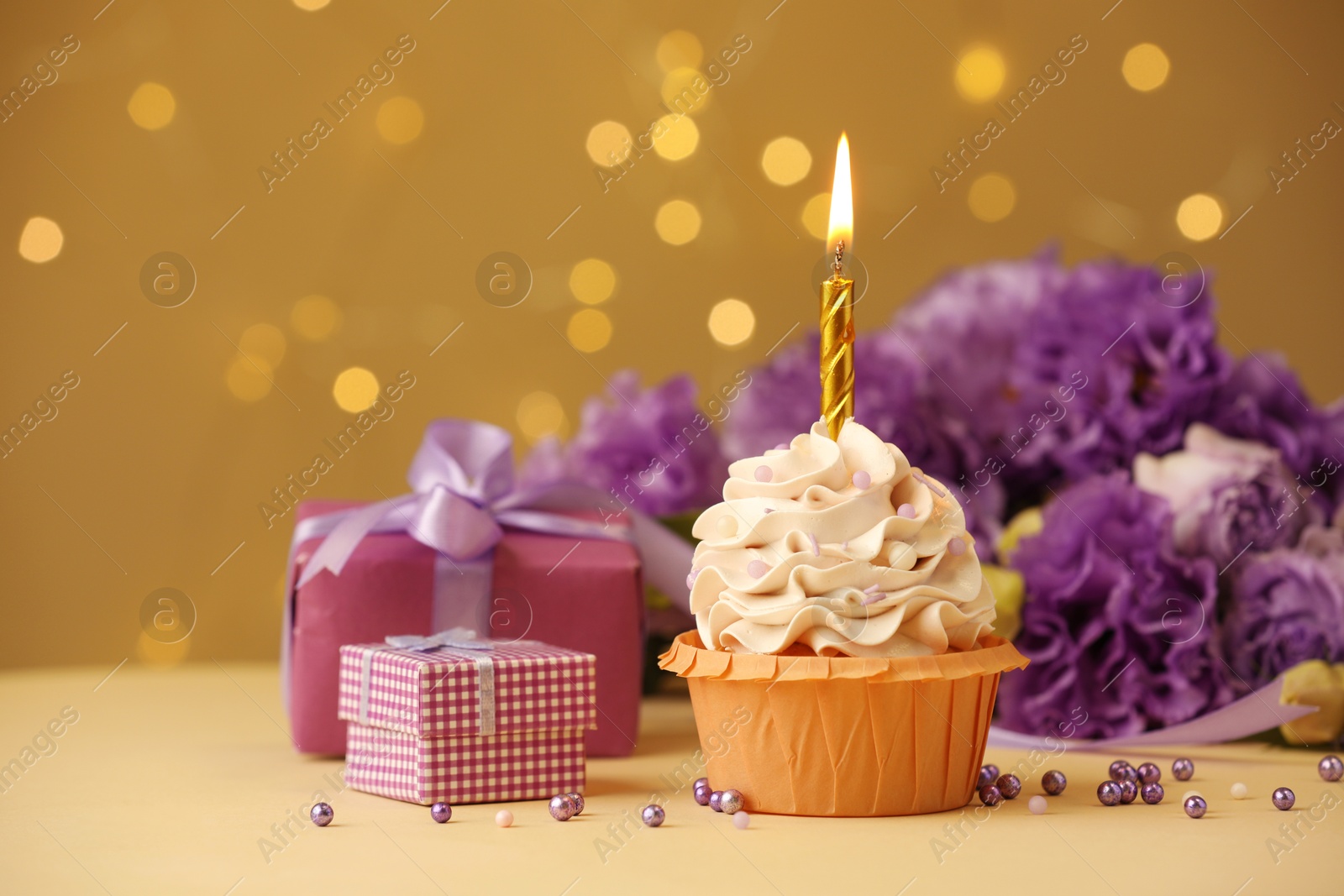 Photo of Birthday cupcake with burning candle, gifts and flowers on beige table against blurred lights, selective focus