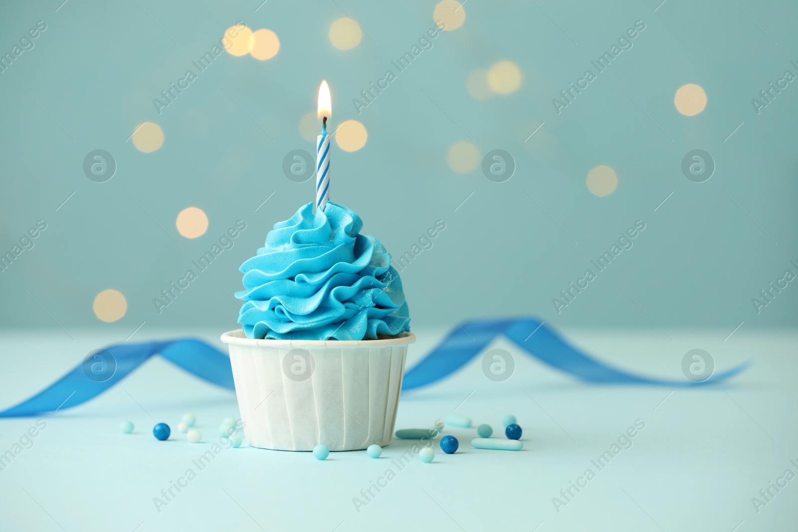 Photo of Birthday cupcake with burning candle on light blue table against blurred lights. Space for text