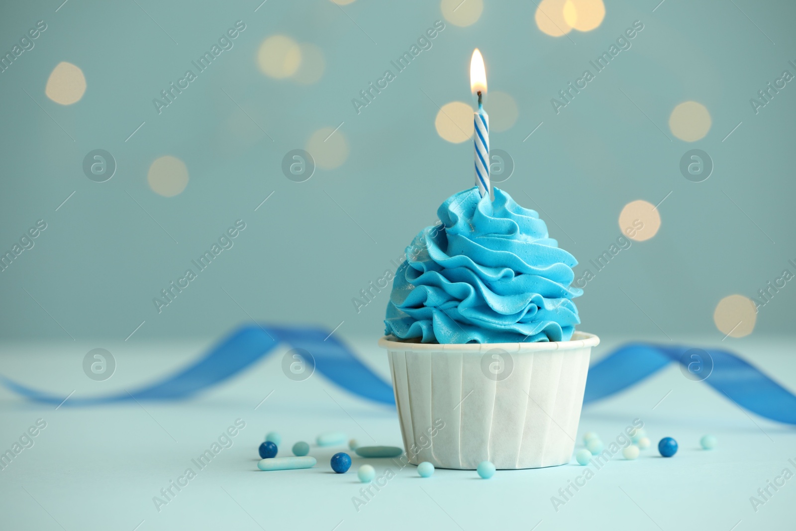 Photo of Birthday cupcake with burning candle on light blue table against blurred lights. Space for text