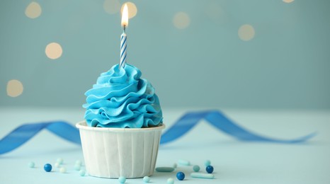 Photo of Birthday cupcake with burning candle on light blue table against blurred lights. Space for text