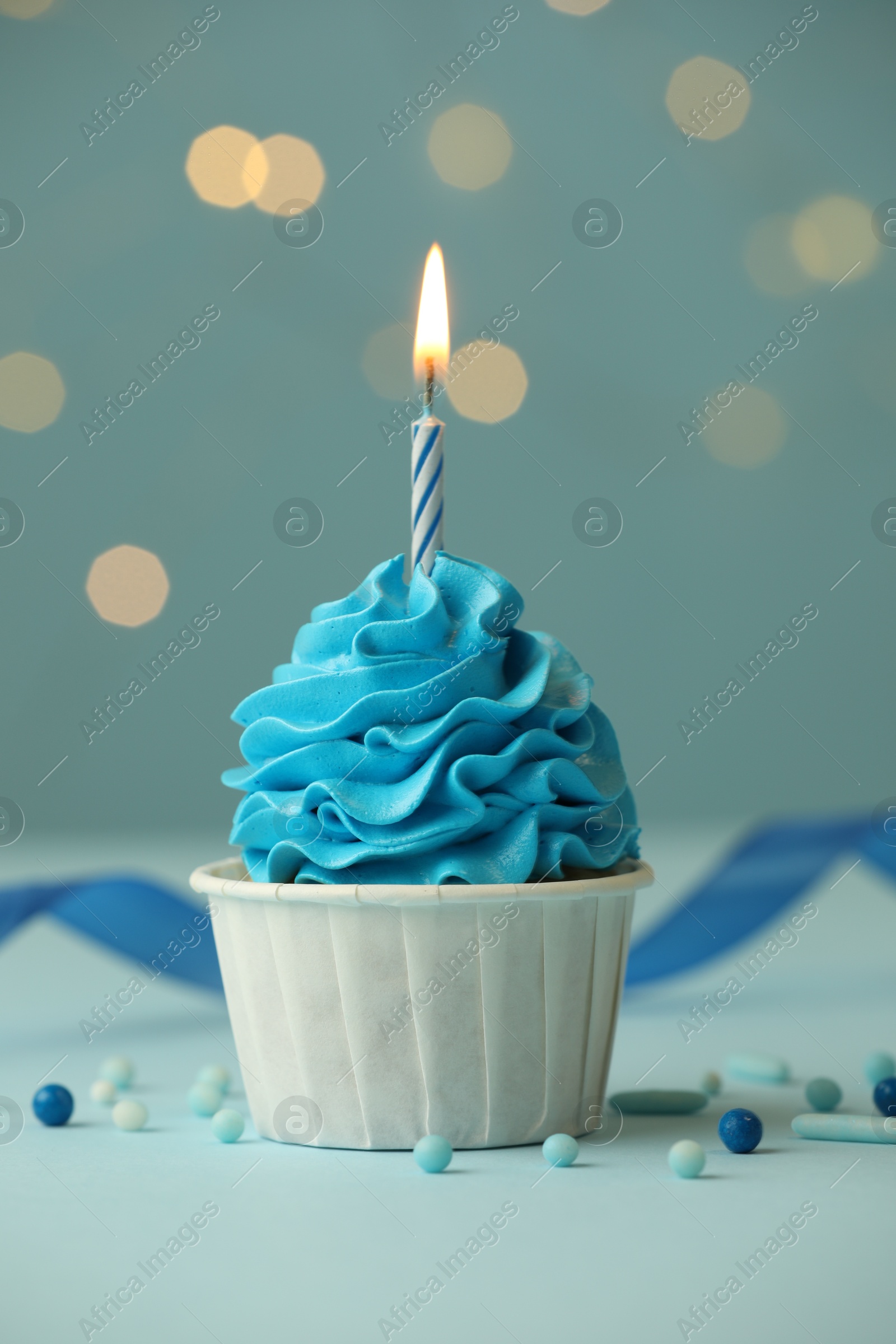 Photo of Birthday cupcake with burning candle on light blue table against blurred lights