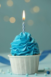 Photo of Birthday cupcake with burning candle on light blue table against blurred lights