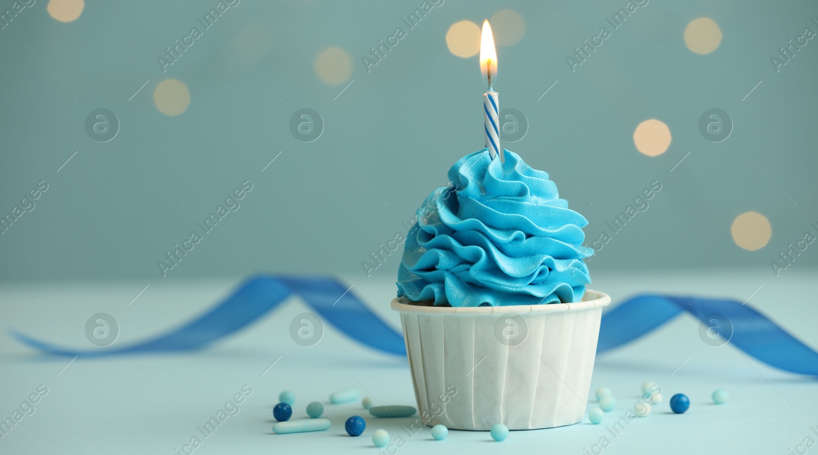 Photo of Birthday cupcake with burning candle on light blue table against blurred lights. Space for text