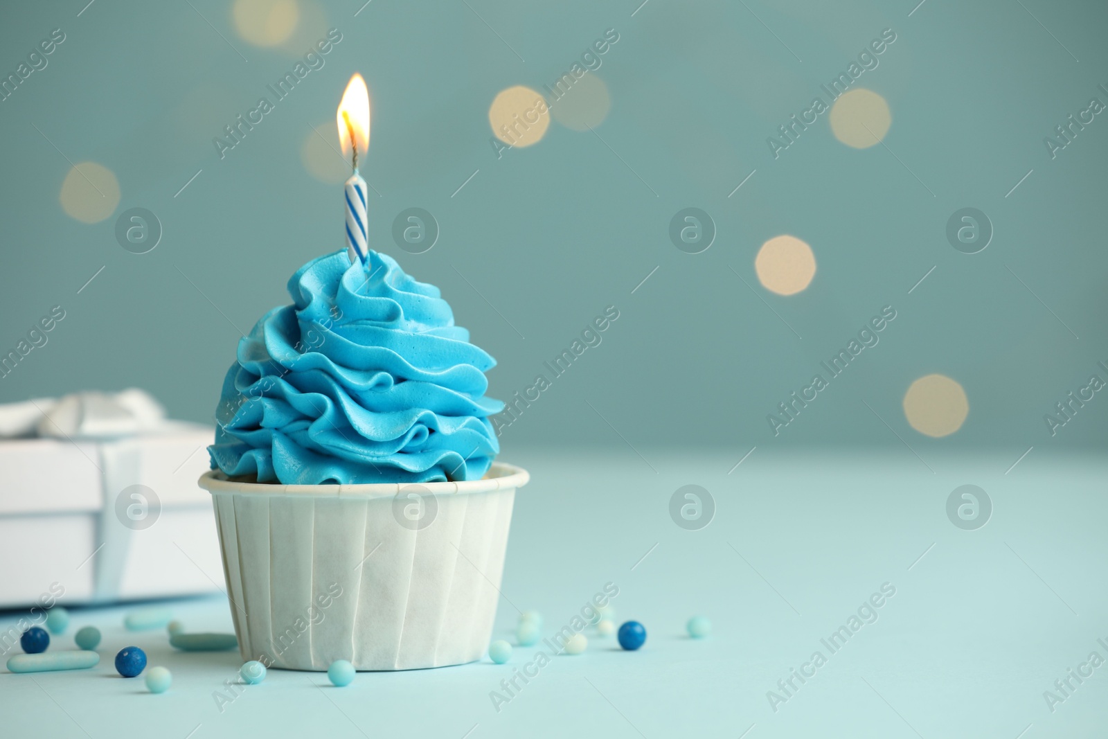 Photo of Birthday cupcake with burning candle and gift on light blue table against blurred lights. Space for text