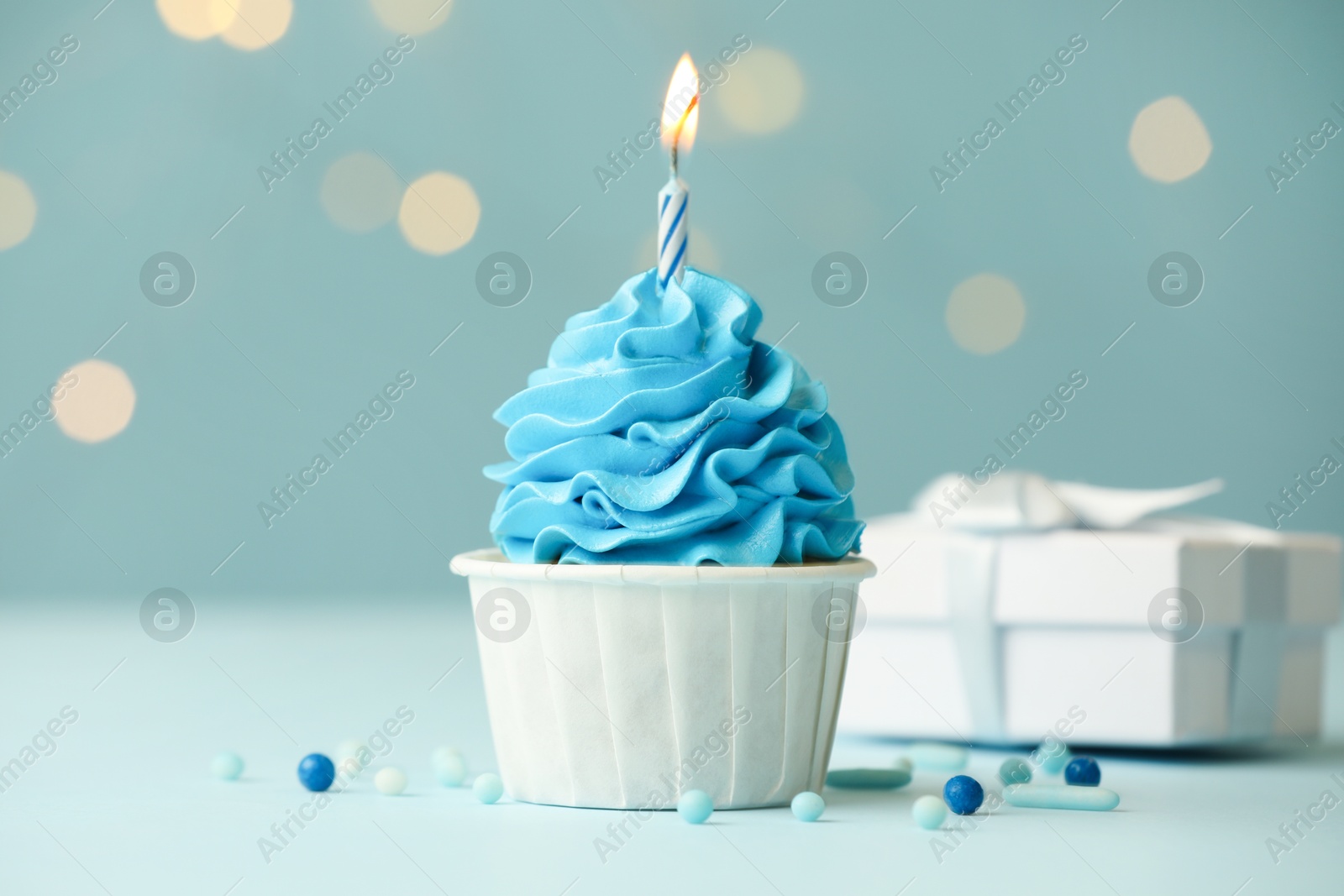 Photo of Birthday cupcake with burning candle and gift on light blue table against blurred lights