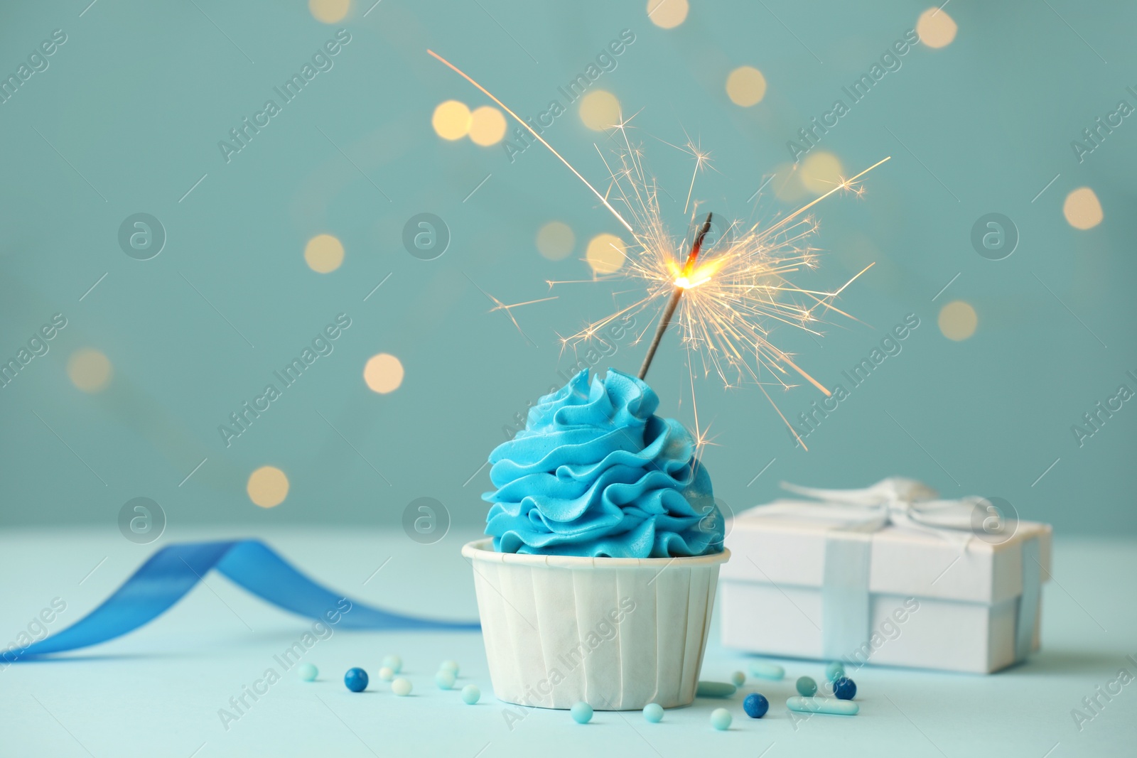 Photo of Birthday cupcake with burning sparkler and gift on light blue table against blurred lights