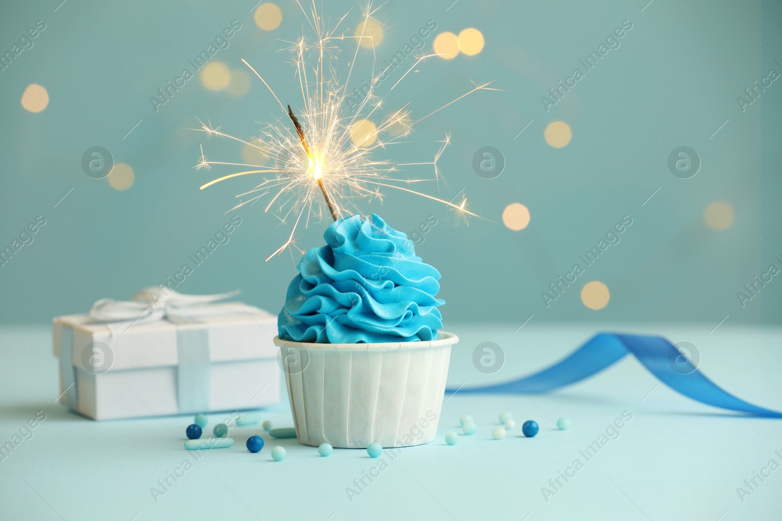 Photo of Birthday cupcake with burning sparkler and gift on light blue table against blurred lights