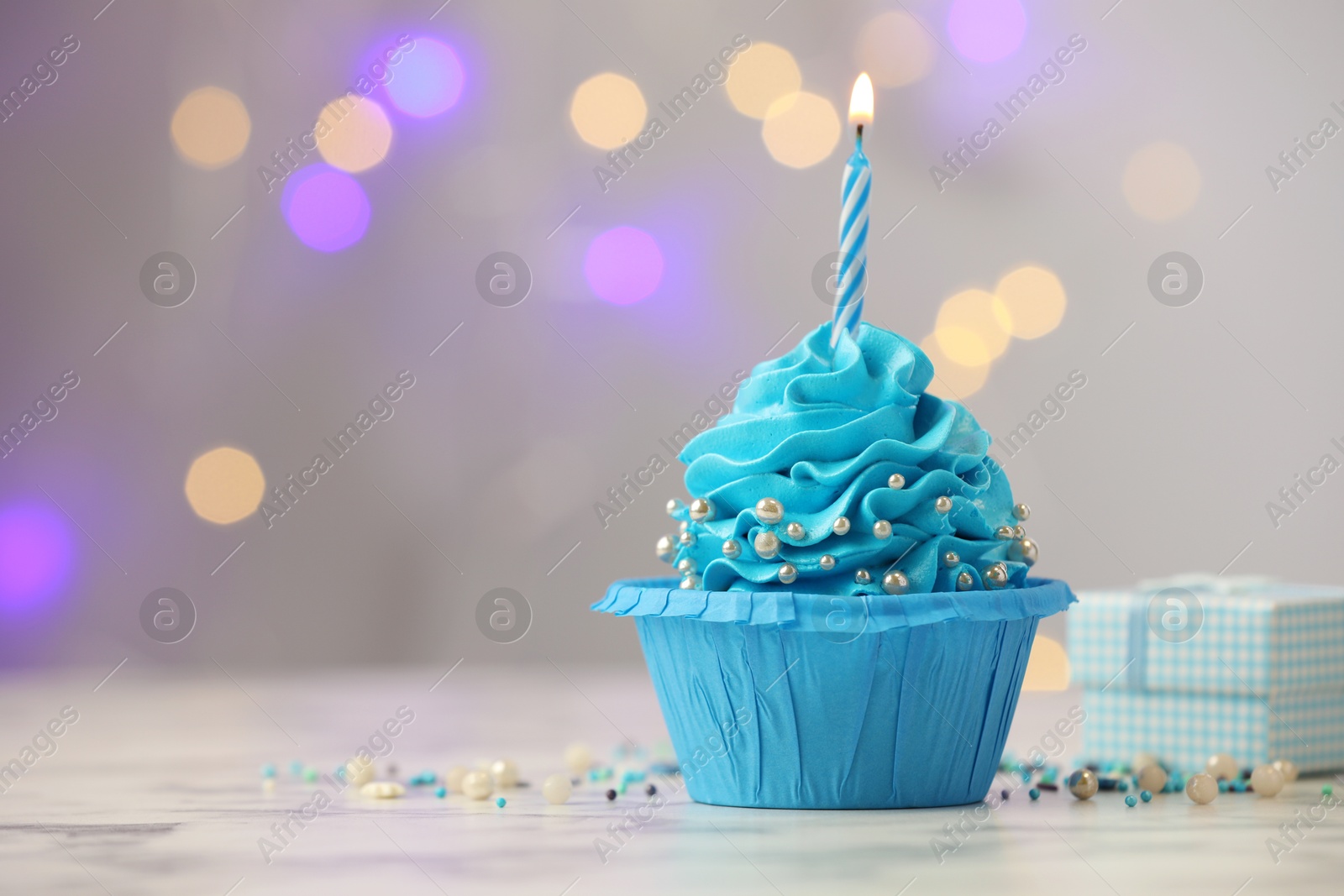 Photo of Birthday cupcake with burning candle and gift on light table against blurred lights. Space for text