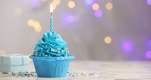 Photo of Birthday cupcake with burning candle and gift on light table against blurred lights. Space for text