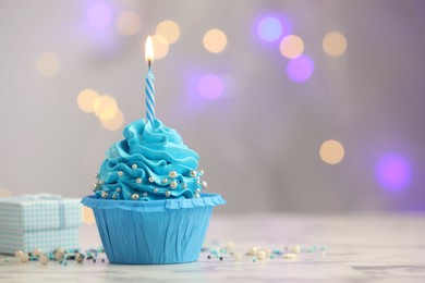 Photo of Birthday cupcake with burning candle and gift on light table against blurred lights. Space for text