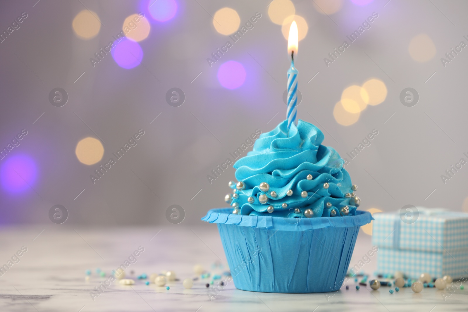 Photo of Birthday cupcake with burning candle and gift on light table against blurred lights. Space for text