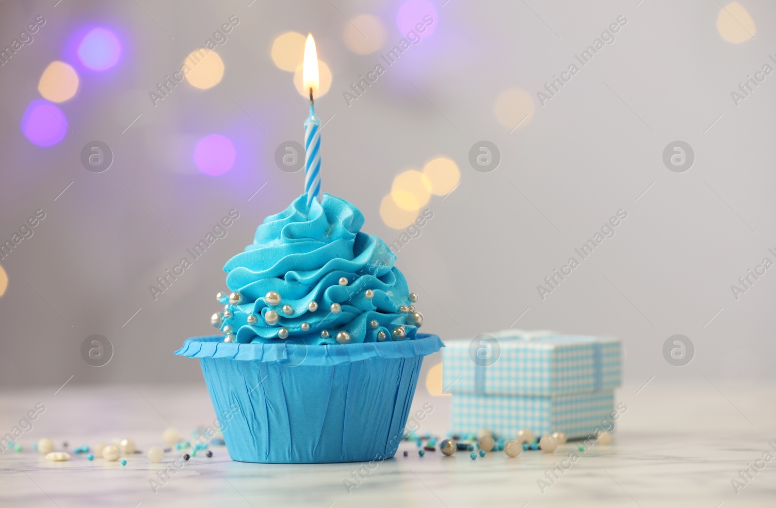 Photo of Birthday cupcake with burning candle and gift on light table against blurred lights