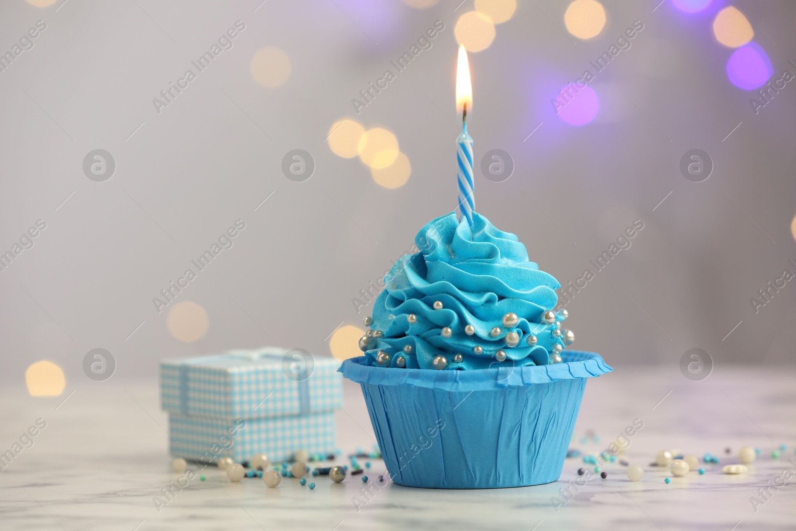 Photo of Birthday cupcake with burning candle and gift on light table against blurred lights