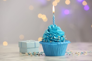 Photo of Birthday cupcake with burning candle and gift on light table against blurred lights