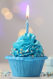 Photo of Birthday cupcake with burning candle on light table against blurred lights