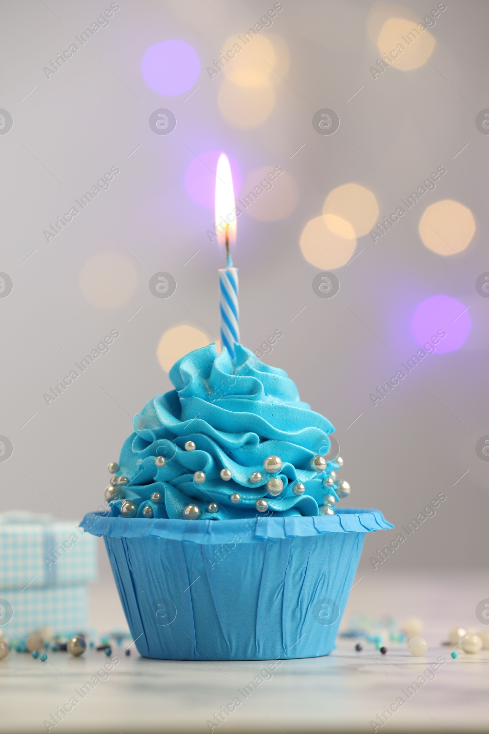 Photo of Birthday cupcake with burning candle on light table against blurred lights