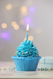 Photo of Birthday cupcake with burning candle on light table against blurred lights