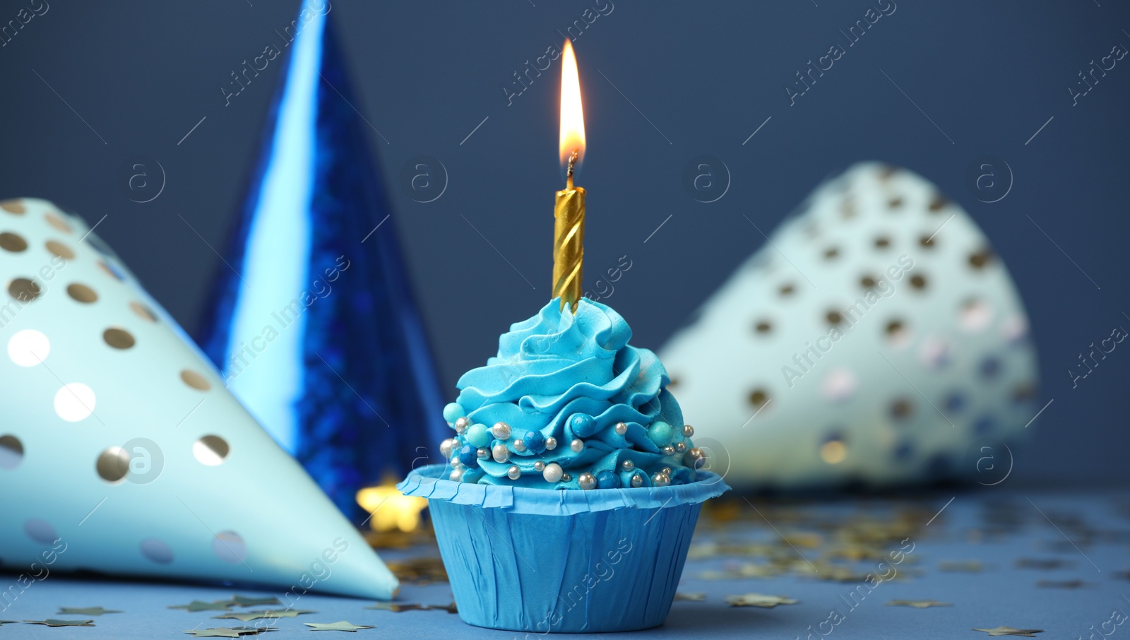 Photo of Birthday cupcake with burning candle and party cones on blue table