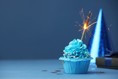 Photo of Birthday cupcake with burning sparkler, gift and party cone on blue table. Space for text