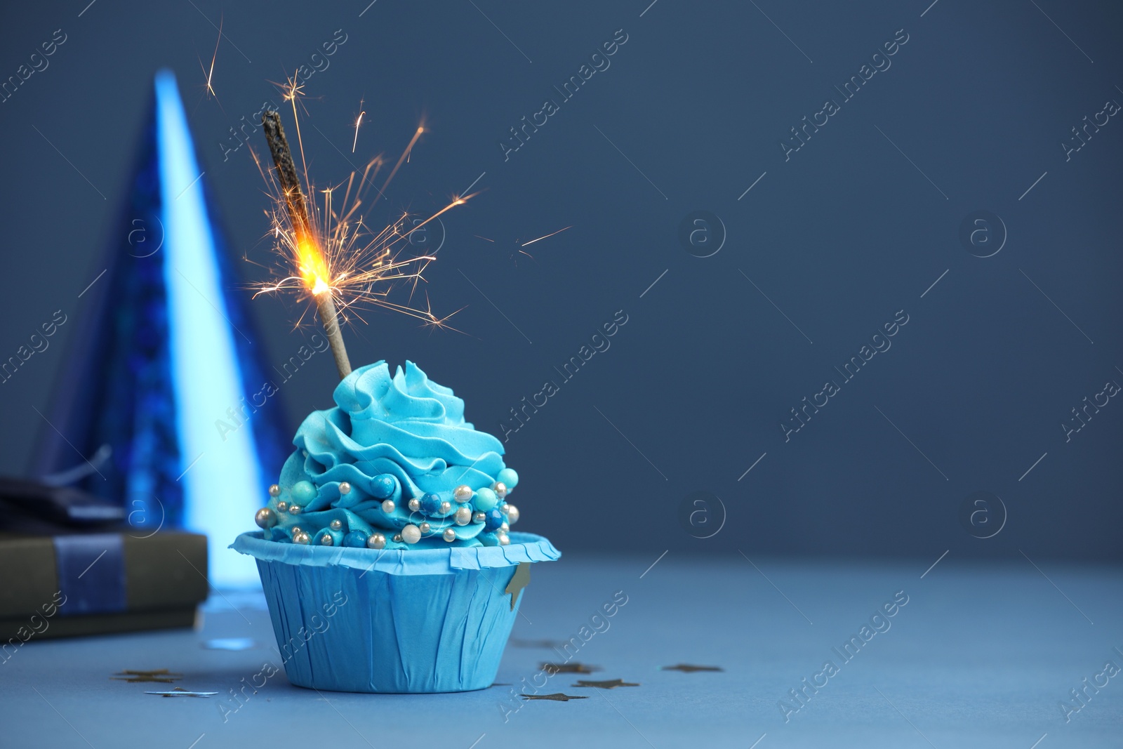 Photo of Birthday cupcake with burning sparkler, gift and party cone on blue table. Space for text