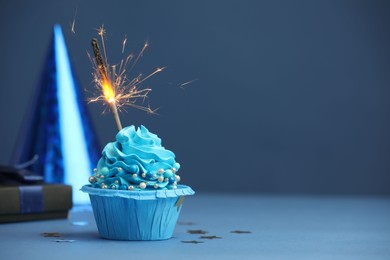 Photo of Birthday cupcake with burning sparkler, gift and party cone on blue table. Space for text