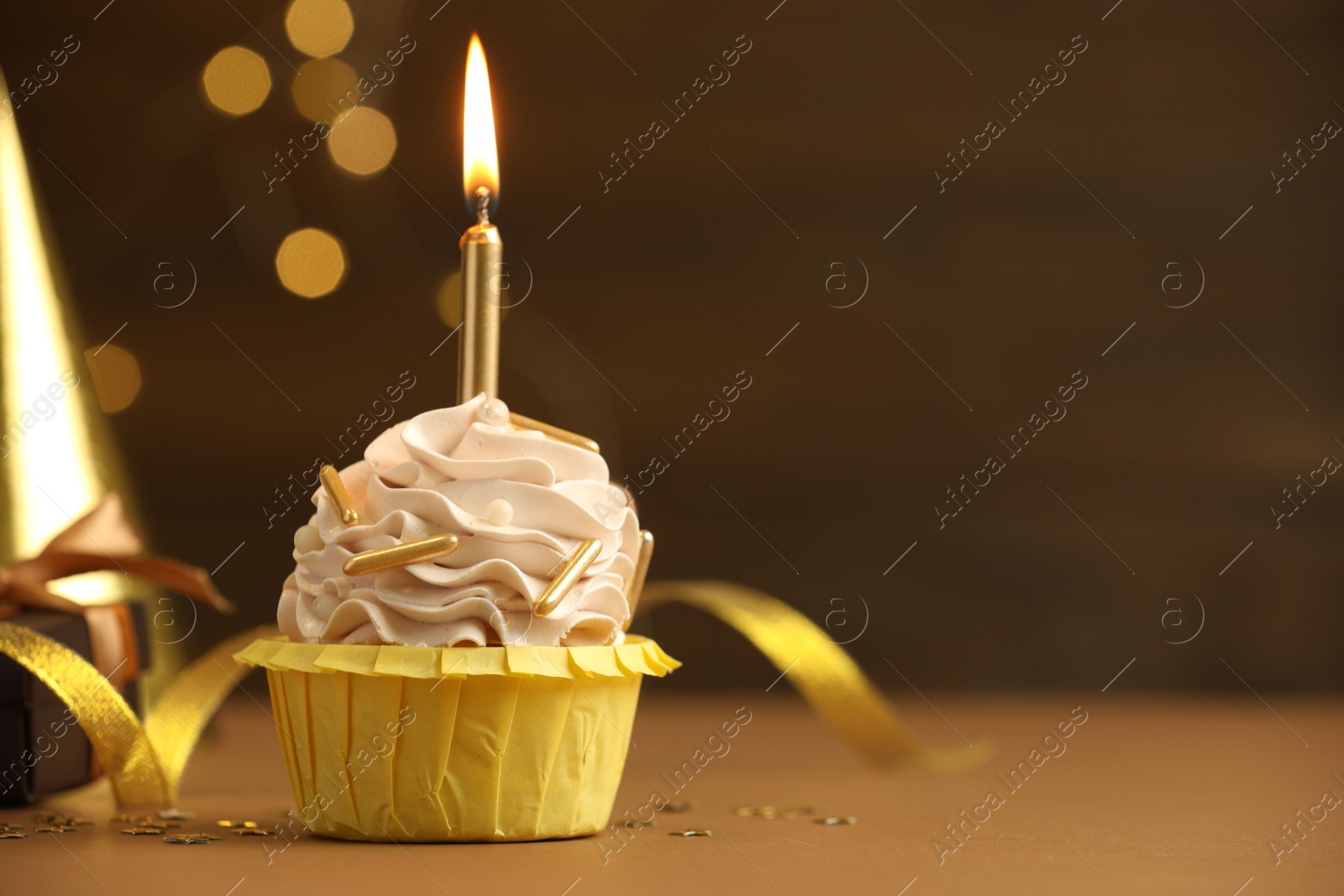 Photo of Birthday cupcake with burning candle on brown table against blurred lights. Space for text