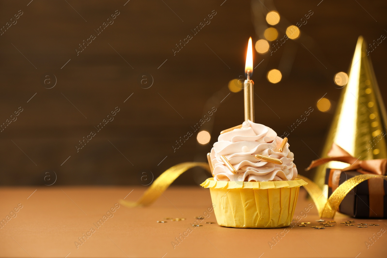 Photo of Birthday cupcake with burning candle on brown table against blurred lights. Space for text