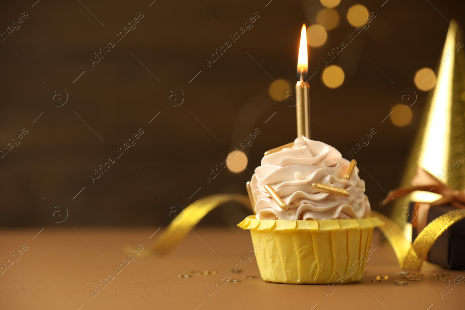 Photo of Birthday cupcake with burning candle on brown table against blurred lights. Space for text