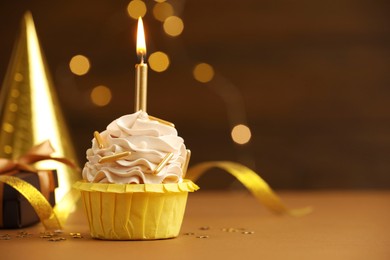 Photo of Birthday cupcake with burning candle on brown table against blurred lights. Space for text