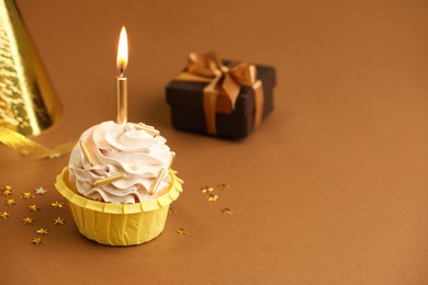 Photo of Birthday cupcake with burning candle, gift and party cone on brown background. Space for text