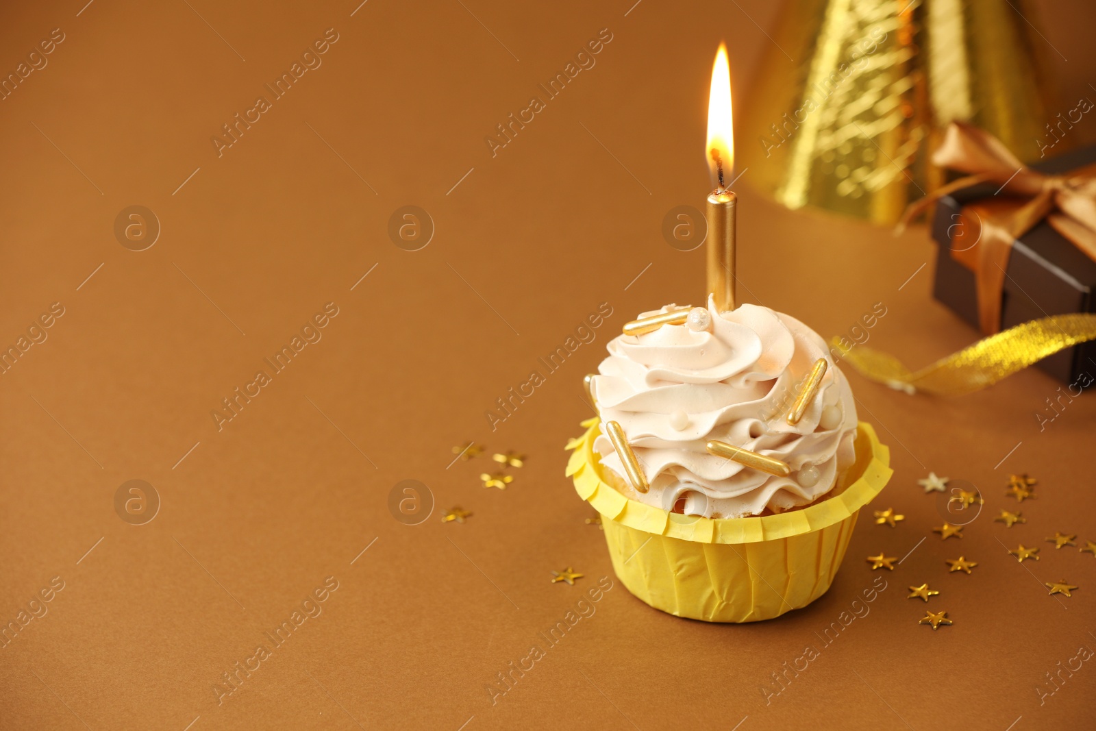 Photo of Birthday cupcake with burning candle, gift and party cone on brown background. Space for text