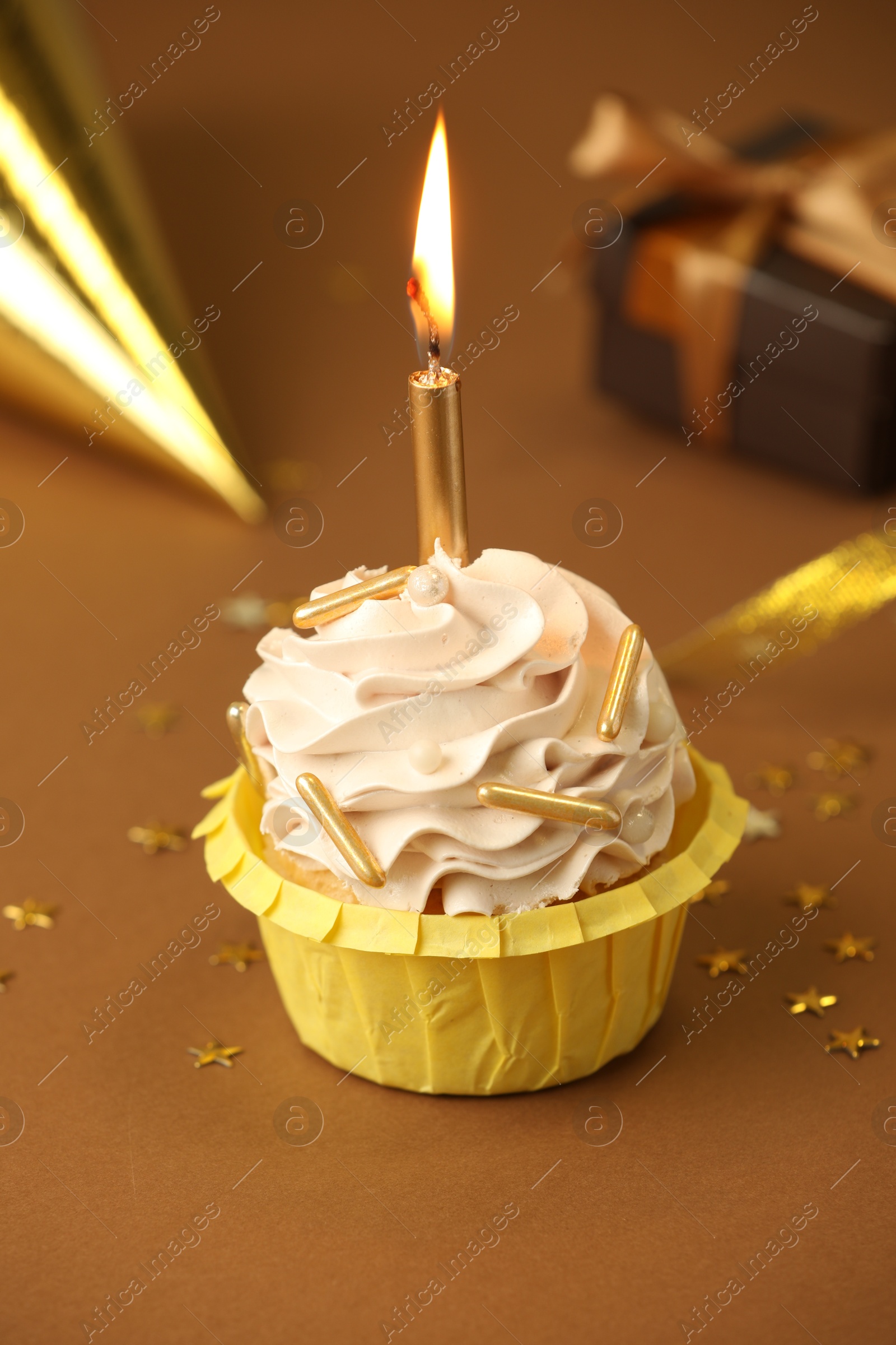 Photo of Birthday cupcake with burning candle, gift and party cone on brown background