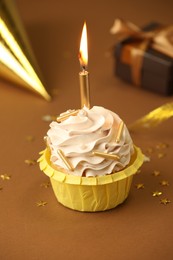 Photo of Birthday cupcake with burning candle, gift and party cone on brown background