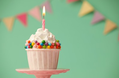 Photo of Birthday cupcake with burning candle on cake stand against turquoise background. Space for text