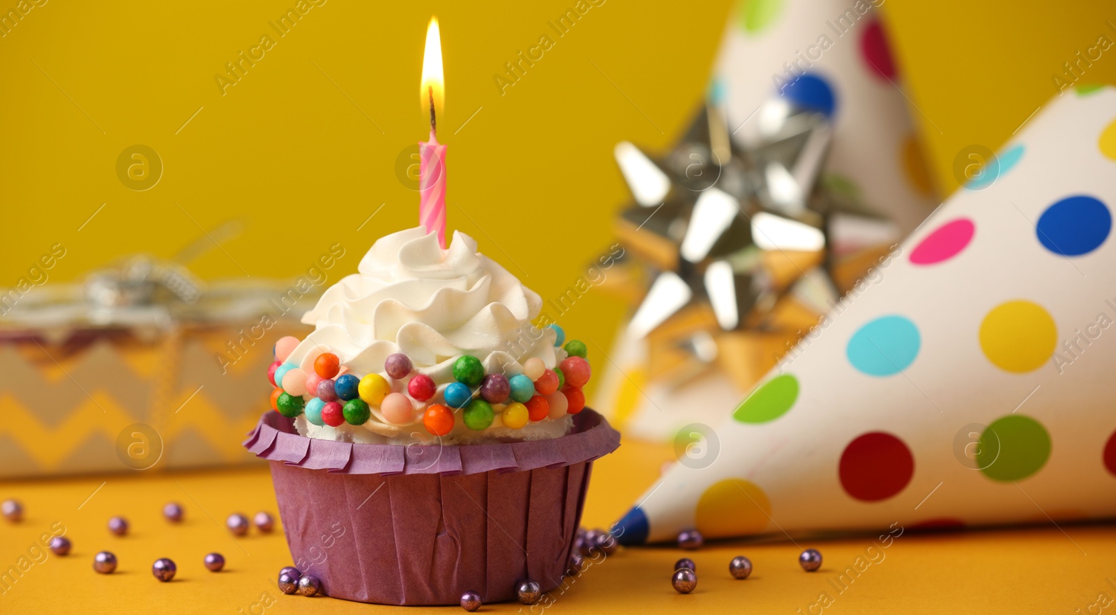 Photo of Birthday cupcake with burning candle and party cone on yellow table