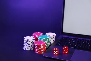 Photo of Online poker. Chips, dice and laptop on dark table, closeup