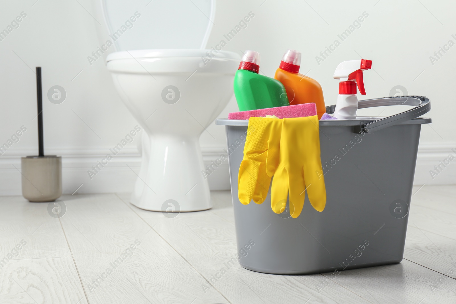 Photo of Bucket with different toilet cleaners and gloves on floor in bathroom